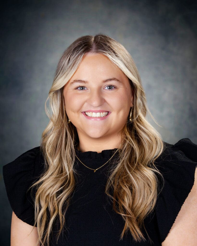 A woman with long, wavy blonde hair, wearing a black top and gold jewelry, smiles at the camera against a dark, softly blurred background.