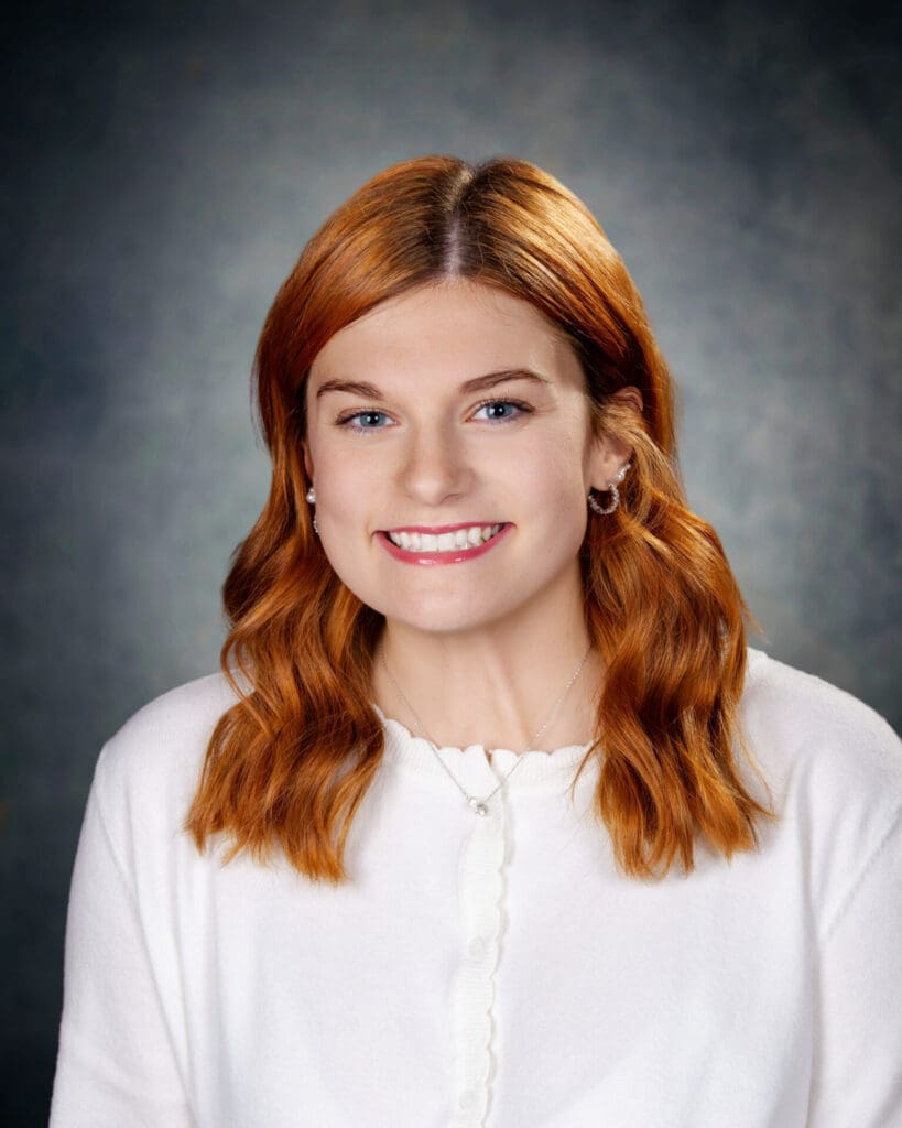 A young woman with wavy red hair, wearing a white sweater and small hoop earrings, smiles at the camera against a dark, softly blurred background.