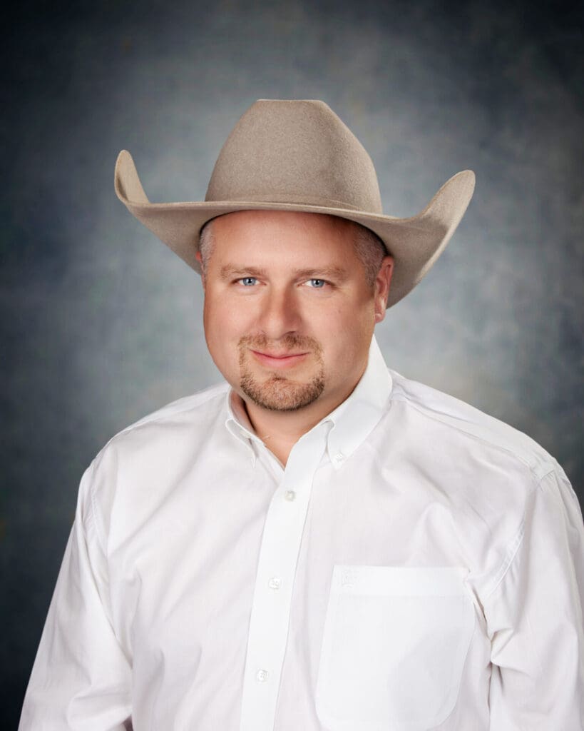 A man wearing a light-colored cowboy hat and a white button-up shirt stands in front of a mottled gray studio background, smiling softly at the camera.