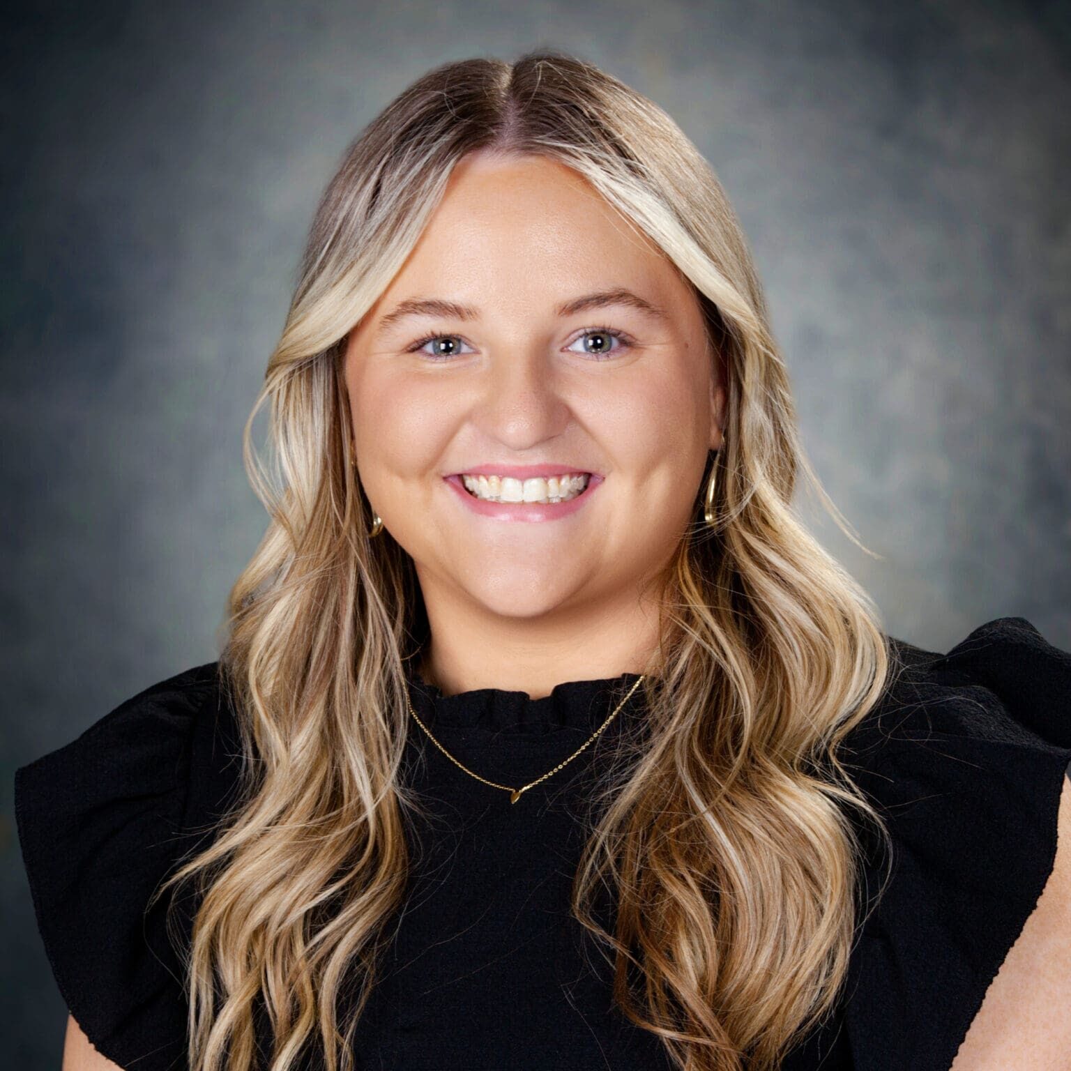 A woman with long, wavy blonde hair, wearing a black top and gold jewelry, smiles at the camera against a dark, softly blurred background.
