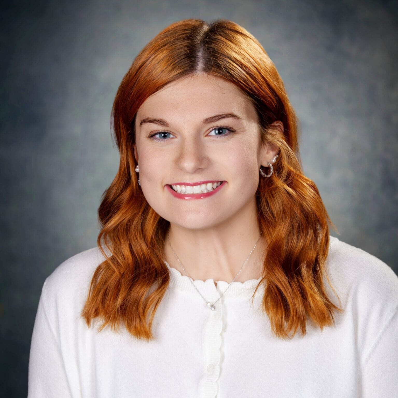 A young woman with wavy red hair, wearing a white sweater and small hoop earrings, smiles at the camera against a dark, softly blurred background.