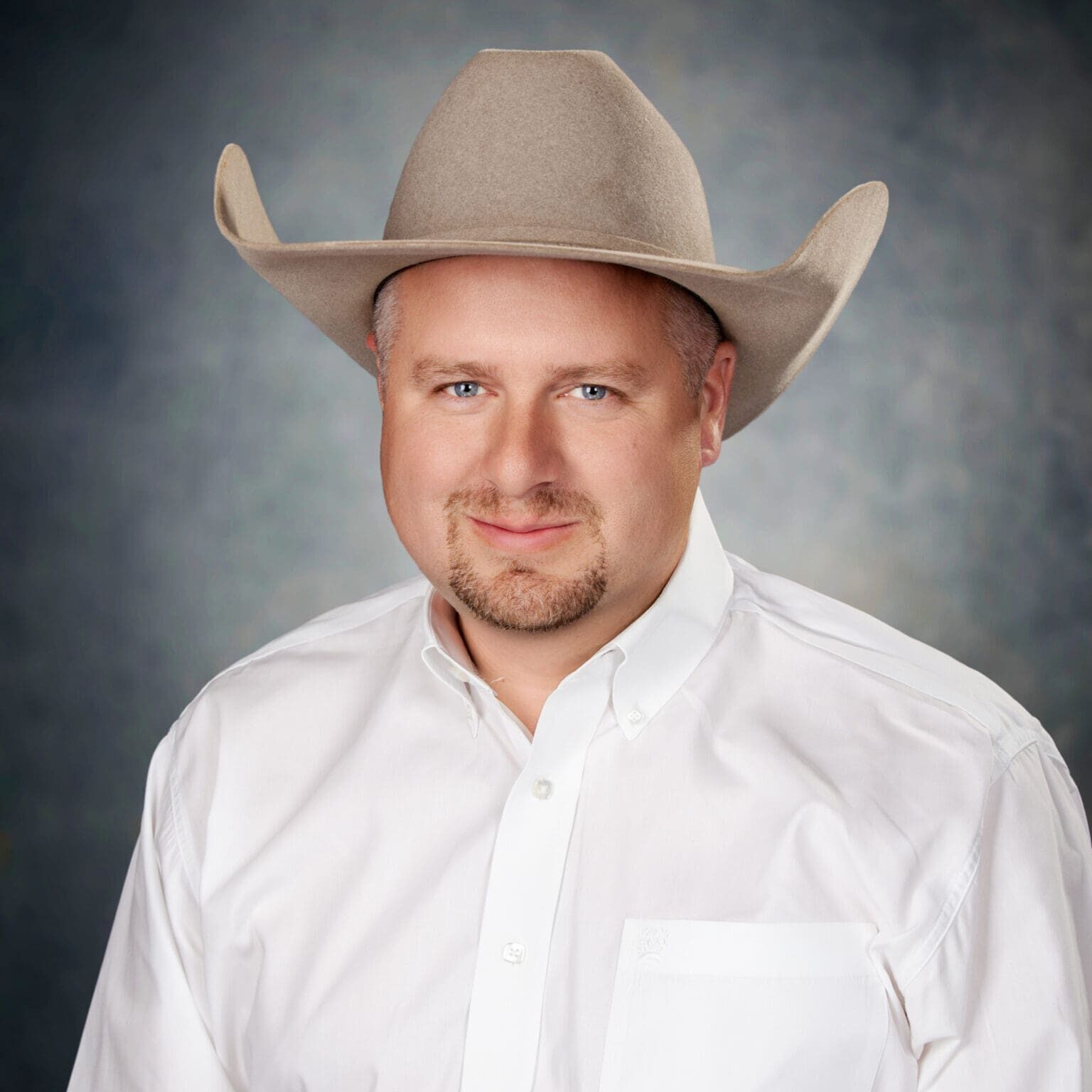 A man wearing a light-colored cowboy hat and a white button-up shirt stands in front of a mottled gray studio background, smiling softly at the camera.