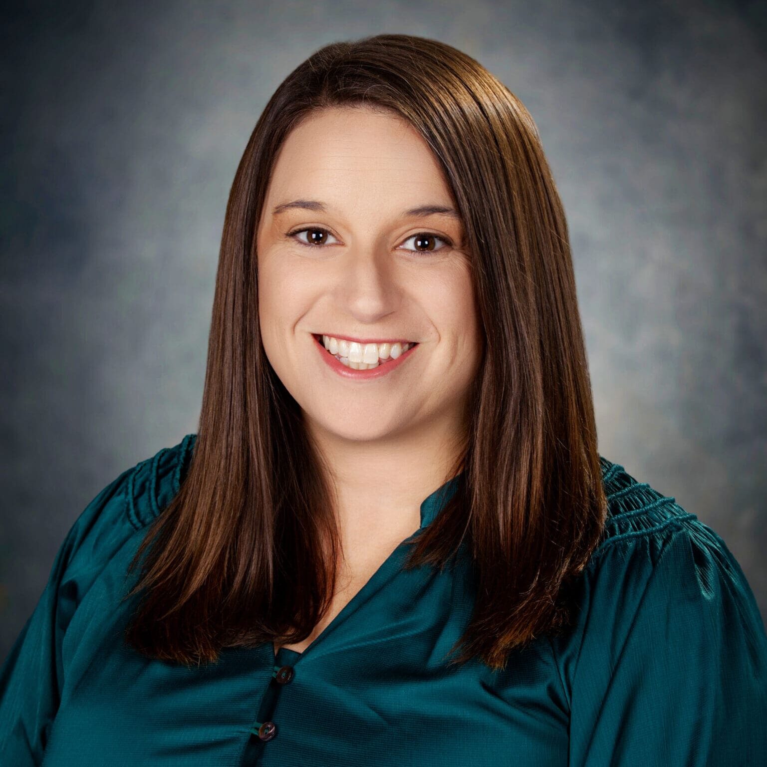 A woman with straight brown hair, wearing a teal blouse with button details, smiles at the camera against a neutral, softly blurred background.
